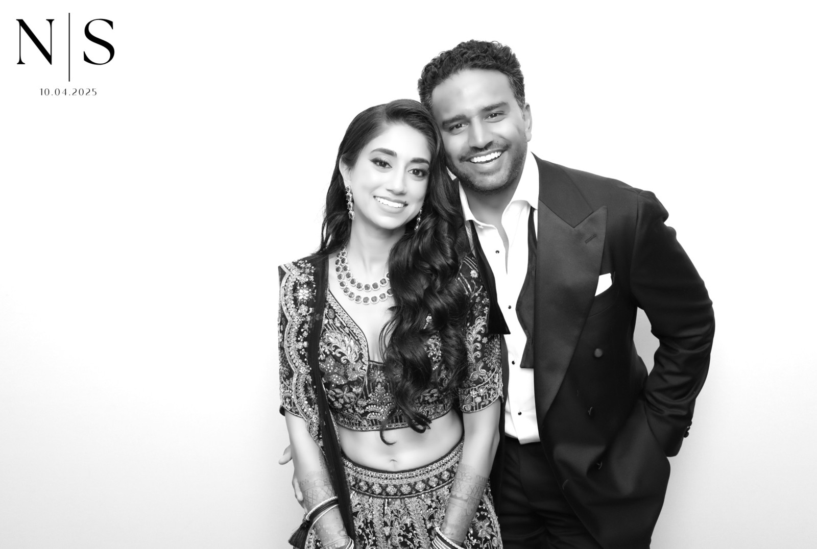 Black-and-white Glam Booth portrait of a South Asian couple, the bride in an embroidered lehenga and the groom in a tuxedo