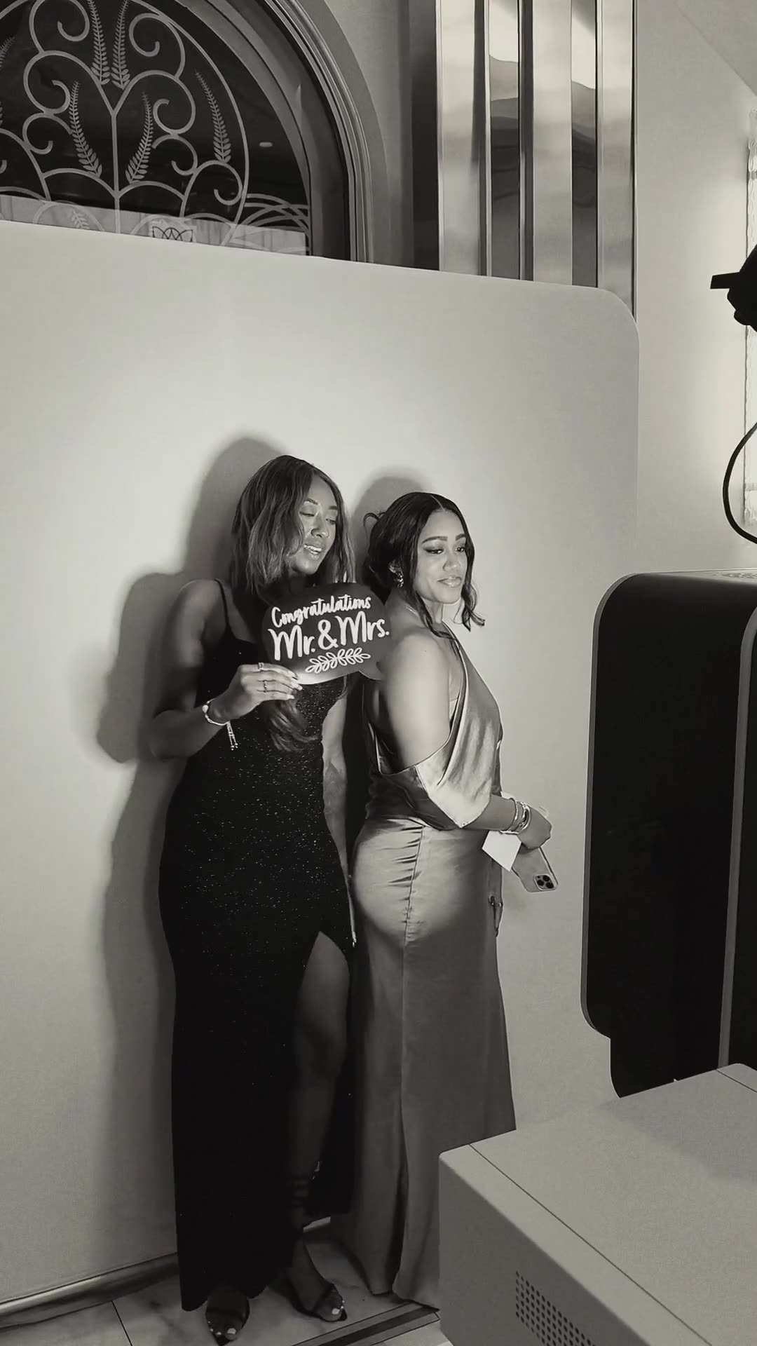 Two guests in evening gowns holding a Congratulations Mr. & Mrs. sign at the black-and-white Glam Booth at a Nigerian wedding reception