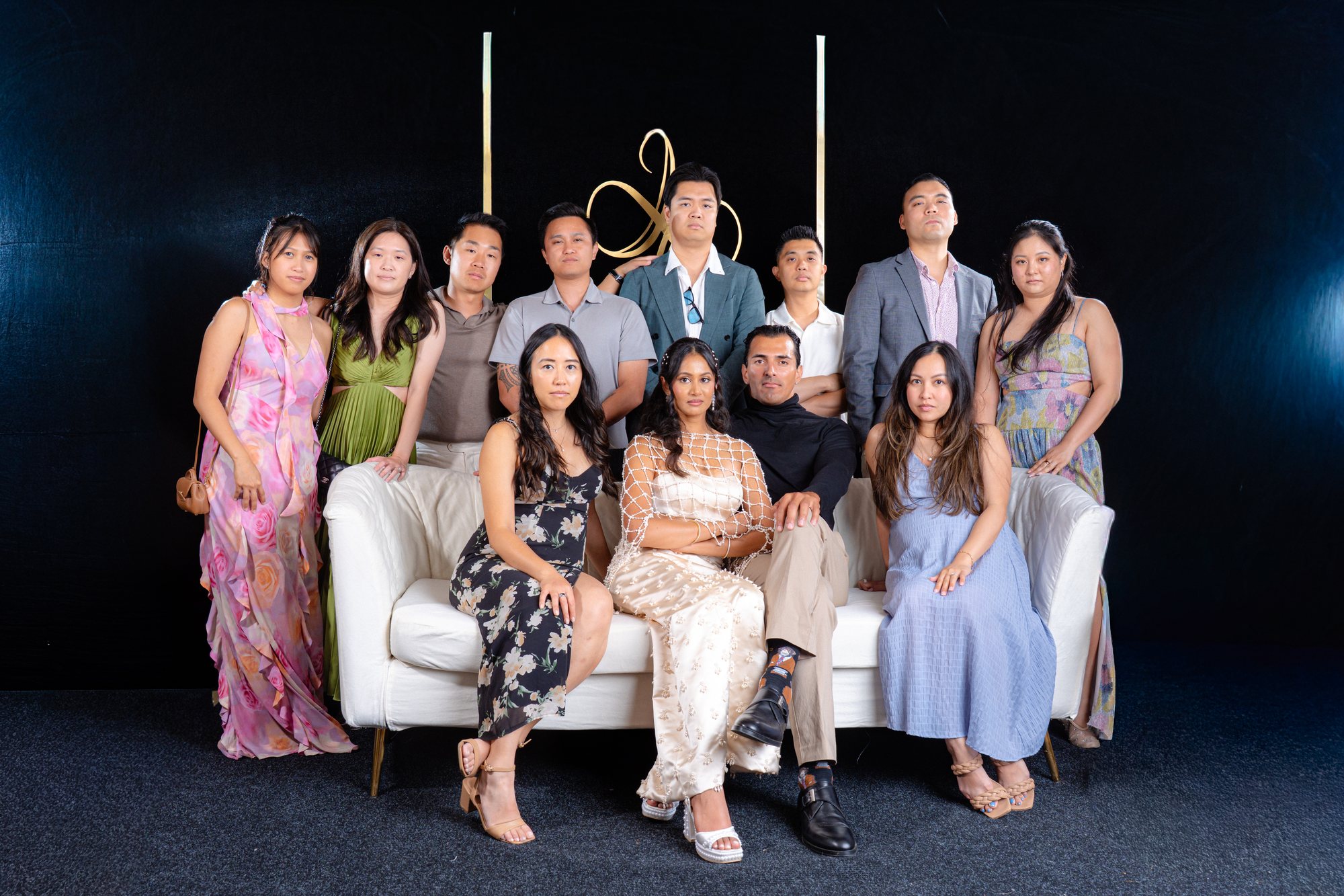Editorial group portrait with twelve guests posed around the white sofa against the black and gold backdrop