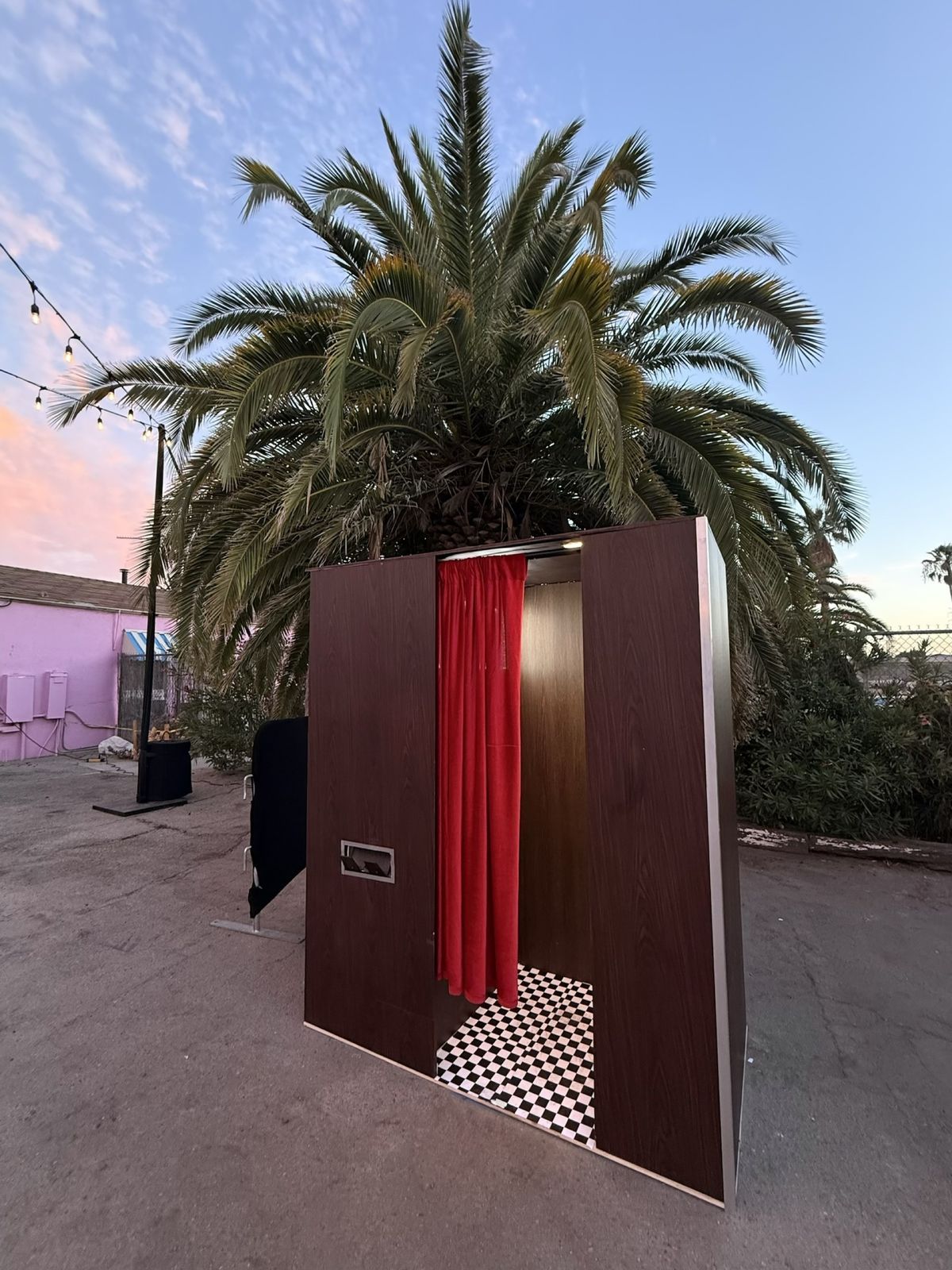 LA Glam Booth enclosed Vintage photo booth with wood-grain walls, red velvet curtain and checkered floor under a palm tree at sunset